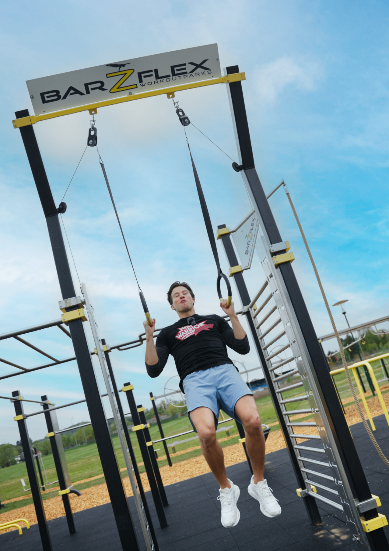 Ein Mann führt Klimmzüge an einem Barz Workout Outdoor-Fitnessstudio durch, trägt ein schwarzes T-Shirt und blaue Shorts.