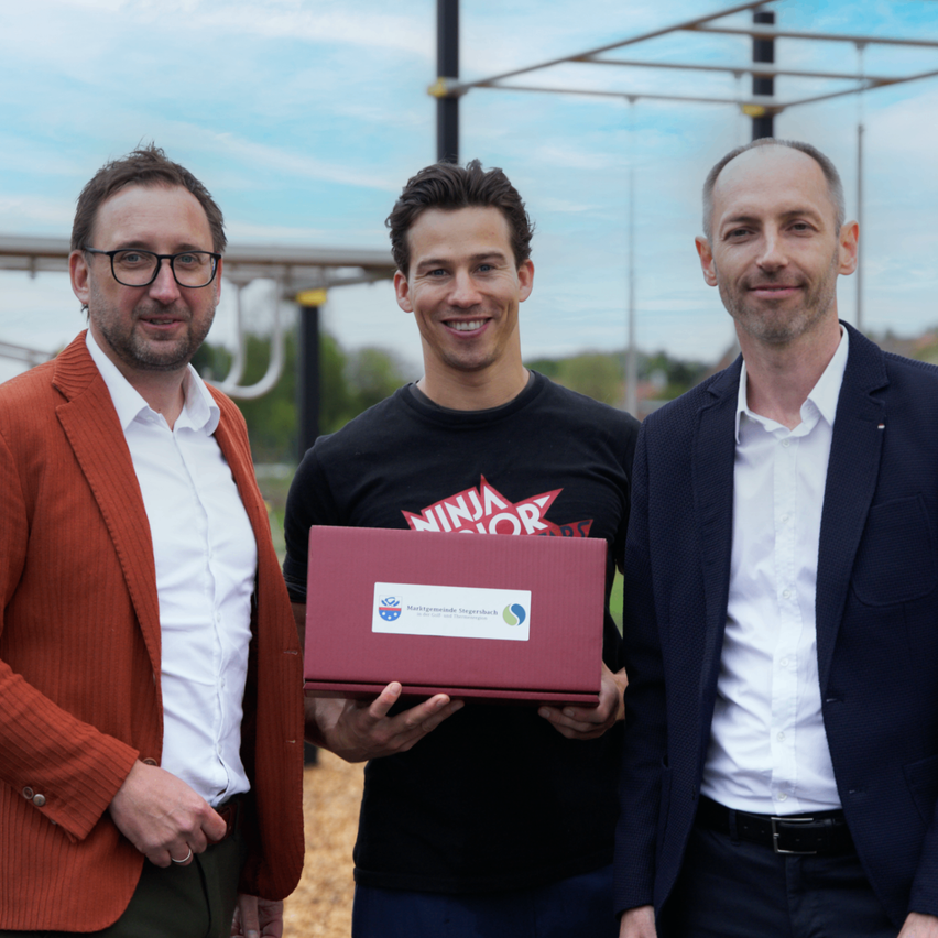 Drei Männer stehen in einem Park und lächeln. Der Mann in der Mitte hält eine rote Box mit einem Logo.