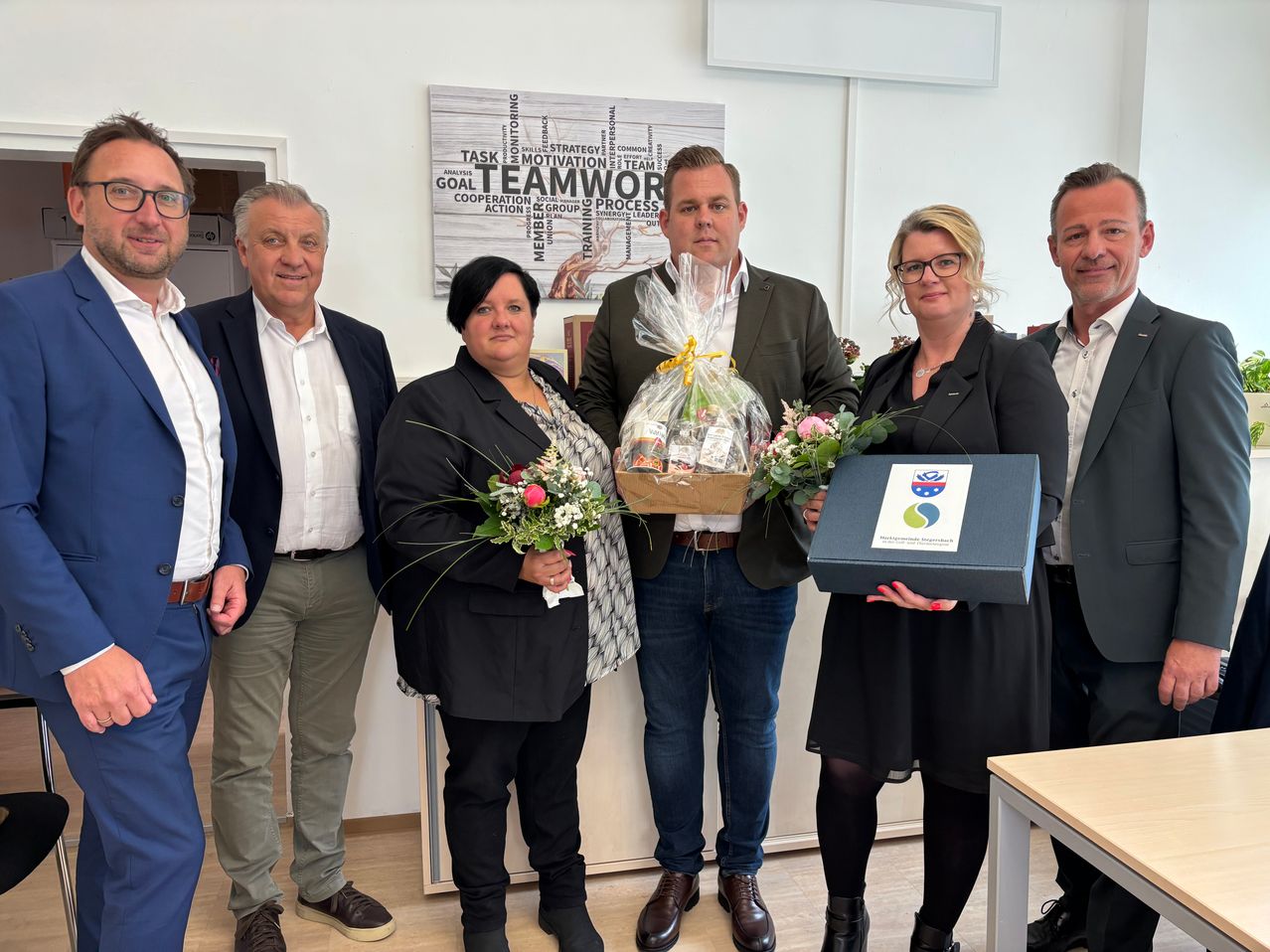 Eine Gruppe von sechs Personen steht in einem Raum und lächelt. Ein Mann und eine Frau halten eine Geschenkbox und einen Blumenstrauß. Dahinter befindet sich eine Wand mit dem Wort Team Work darauf.