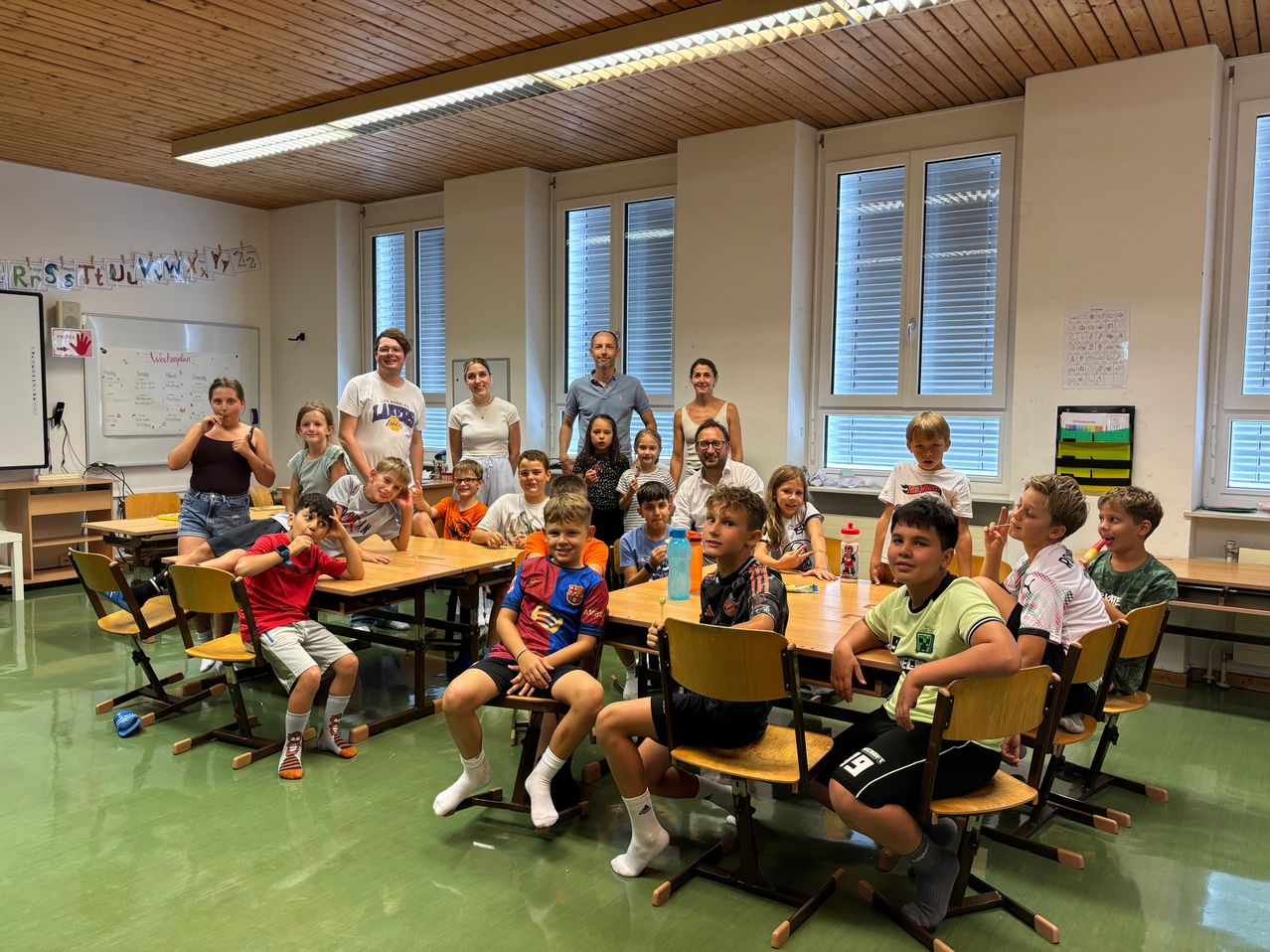 Eine Gruppe von Kindern und Erwachsenen befindet sich in einem Klassenzimmer, einige sitzen an Tischen, andere stehen, alle lächeln für ein Foto. Eine Tafel und Bücher sind im Hintergrund zu sehen.