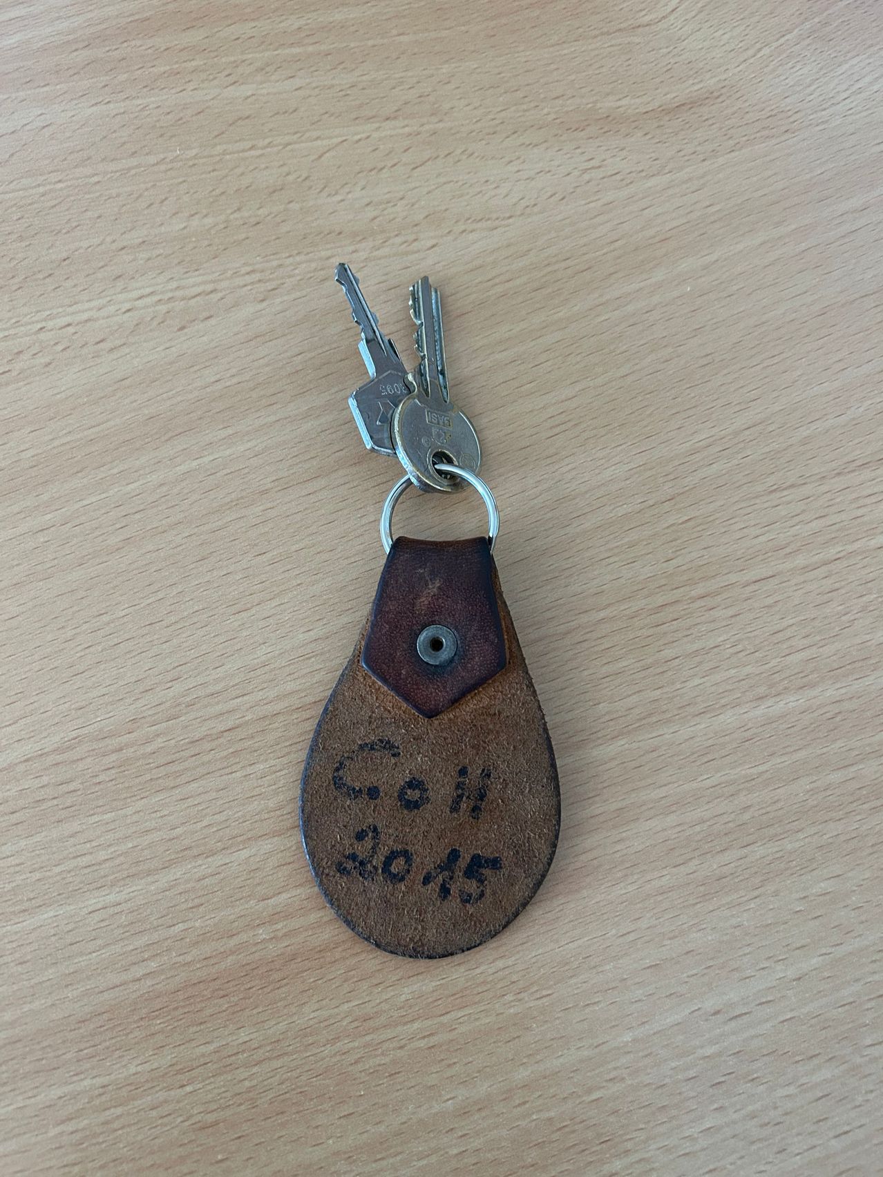 Ein Schlüsselanhänger mit zwei Schlüsseln, der einen braunen Lederanhänger mit 'Coh' und '2015' graviert hat. Der Schlüsselanhänger liegt auf einer Holzoberfläche.