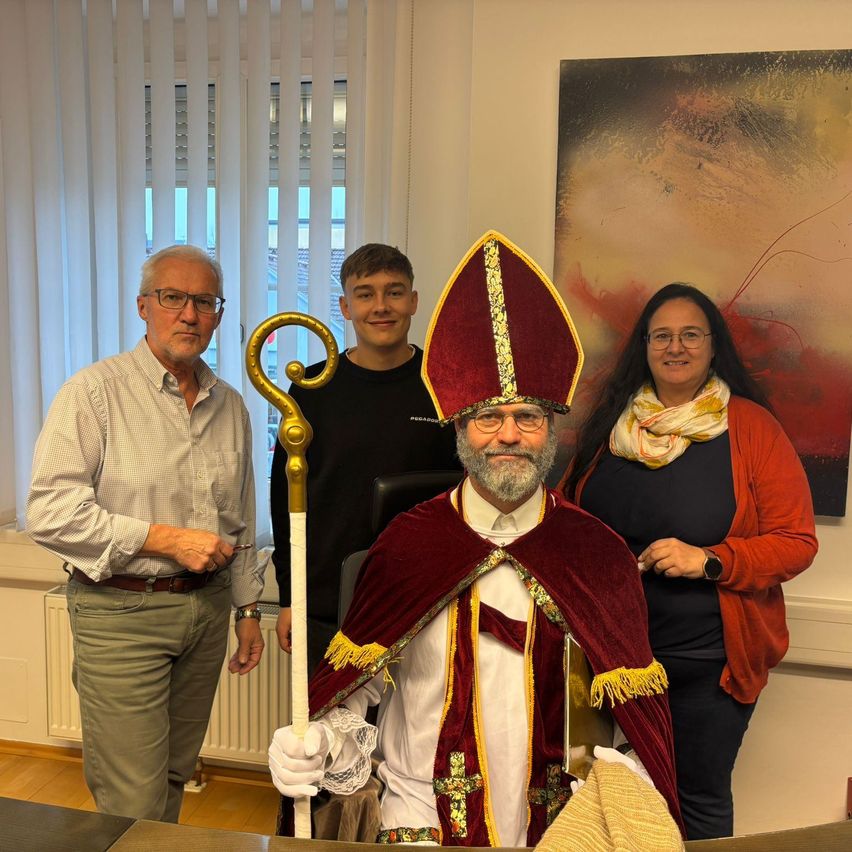 Drei Personen in einem Raum mit einem Mann in einem roten und goldenen Gewand, lächelnd, möglicherweise für ein Foto.