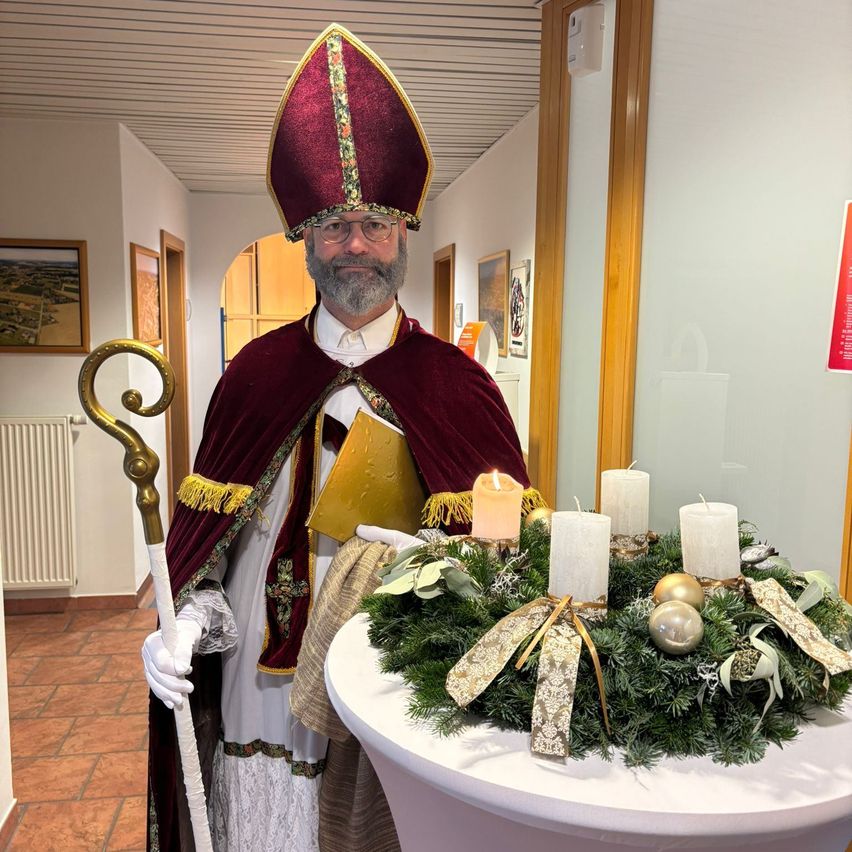 Ein Mann, der als Bischof gekleidet ist, steht neben einem Tisch mit einer Girlande und vier brennenden Kerzen. Er hält einen Bischofsstab. Der Raum hat weiße Wände und geflieste Böden.