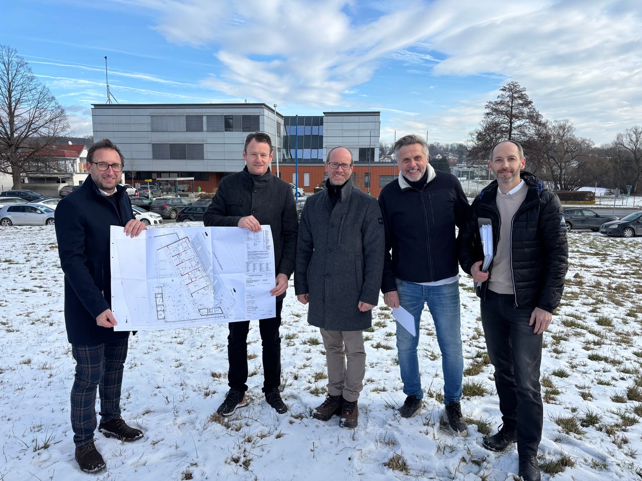 Fünf Männer in Winterkleidung stehen im Schnee, einer hält einen Bauplan. Hinter ihnen sind Gebäude und geparkte Autos unter einem bewölkten Himmel zu sehen.