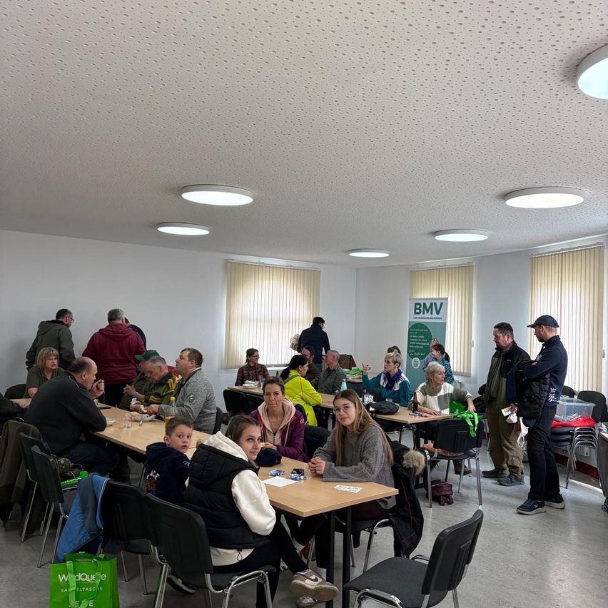 Mehrere Personen sitzen an Tischen in einem Raum, einige stehen herum. An der Wand ist ein Banner mit der Aufschrift BMV zu sehen. Einige Tische haben Essen und Getränke. Der Raum hat weiße Wände, Fenster mit Jalousien und eine Decke mit Lichtern.