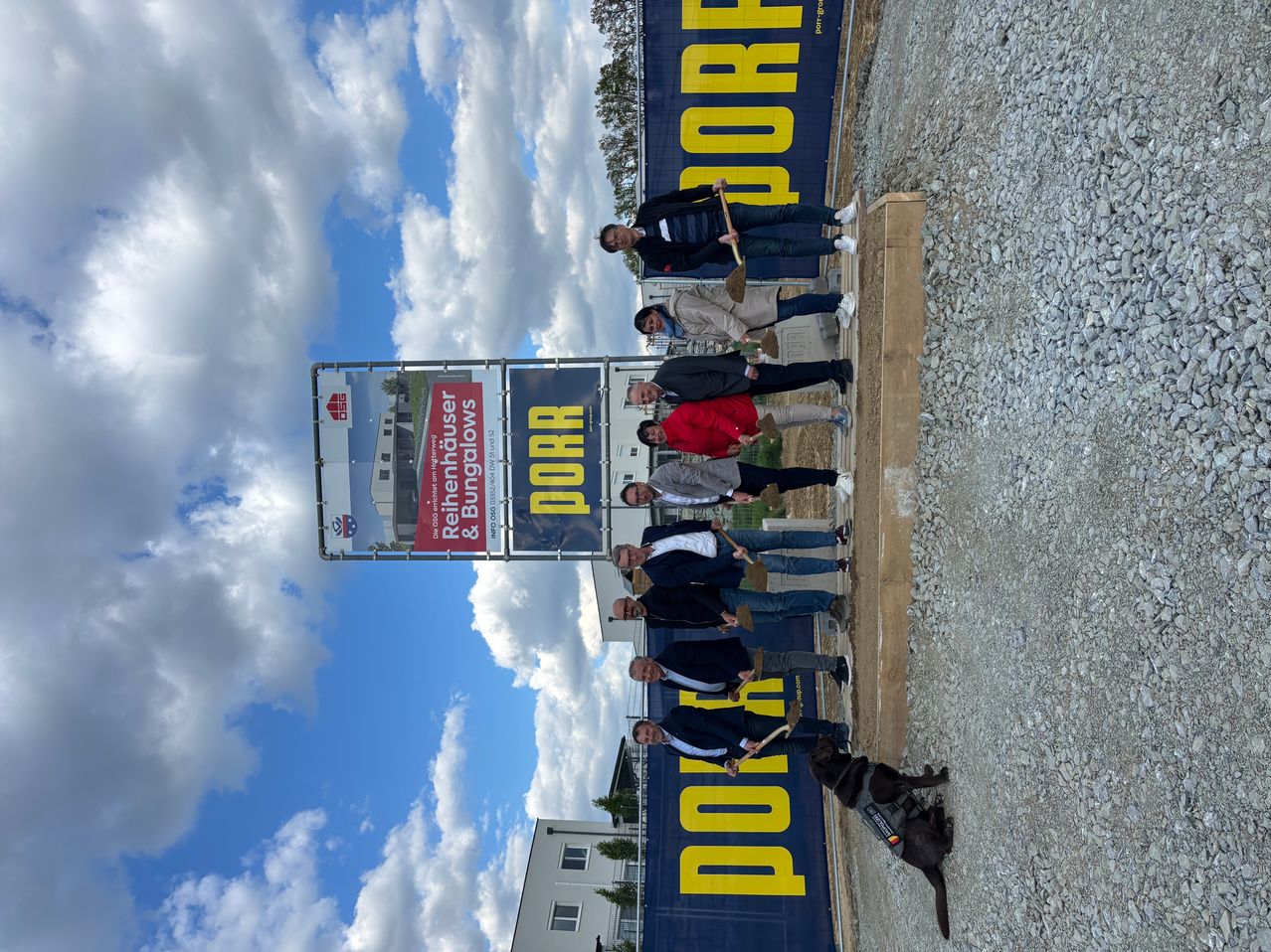 Eine Gruppe von Menschen in formeller Kleidung posiert für ein Foto an einer Baustelle. Sie halten Schaufeln und stehen vor einem blau-gelben Banner. Der Himmel ist blau mit weißen Wolken.