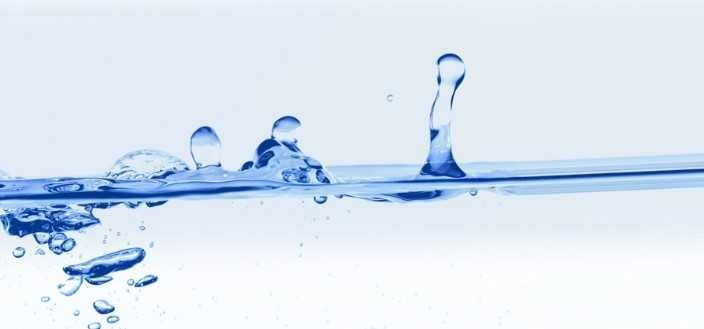 Eine Nahaufnahme von Wassertropfen, die in eine klare blaue Flüssigkeit fallen und dabei Wellen und Blasen auf der Oberfläche erzeugen.