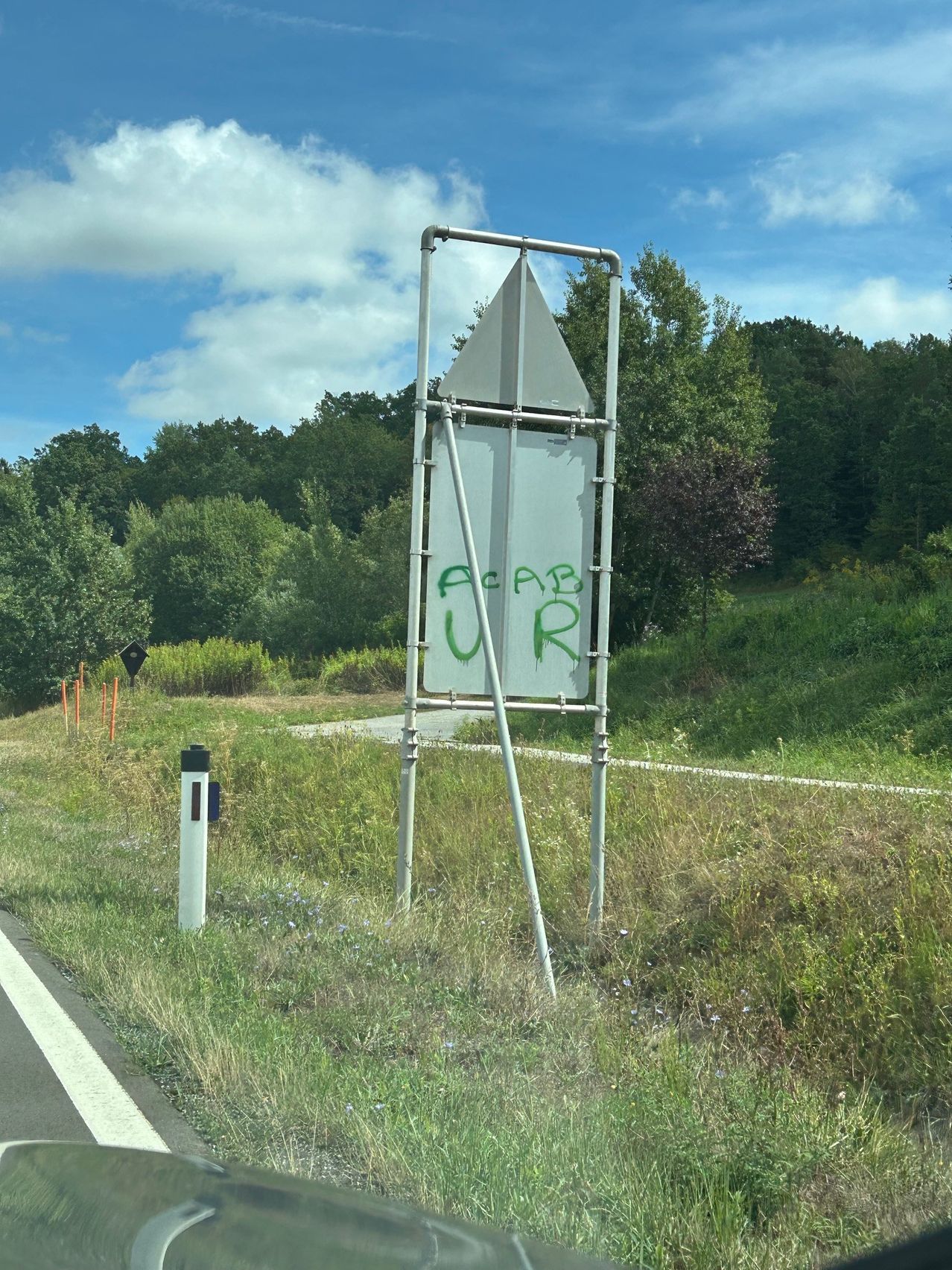 Ein beschädigtes Straßenschild mit grüner Graffiti, die 'AcpB UR' liest, steht am Straßenrand in der Nähe eines Grasbereichs mit Bäumen.
