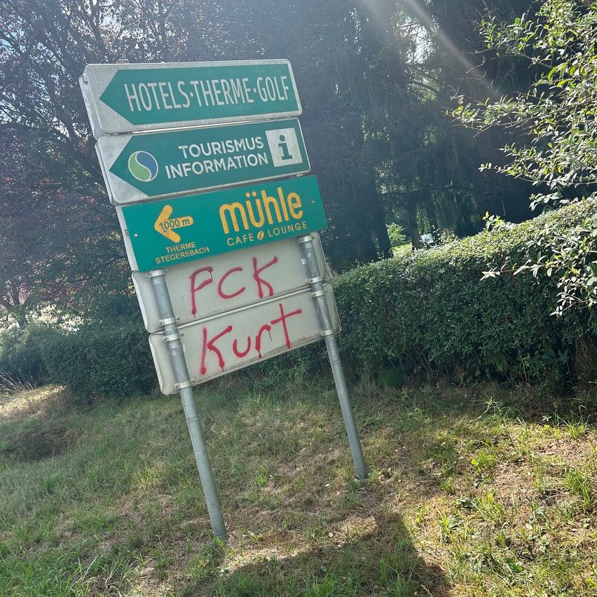 Ein Schild mit mehreren Schildern, die auf Hotels, Golf und Tourismusinformation hinweisen, mit einer roten Graffiti, die 'FCK Kurt' auf dem unteren Schild steht.