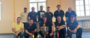 A group of men, likely colleagues, pose for a photo with trophies in front of an arched window. Some wear hats and glasses.