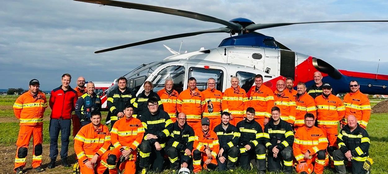 Eine Gruppe Feuerwehrleute in orangefarbenen Uniformen posiert vor einem Hubschrauber unter einem bewölkten Himmel.