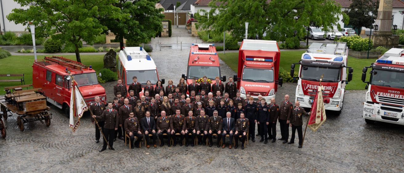 Eine Gruppe Feuerwehrleute in Uniform steht vor Feuerwehrwagen und Fahnen auf einem Kopfsteinpflaster mit Häusern und Bäumen im Hintergrund.