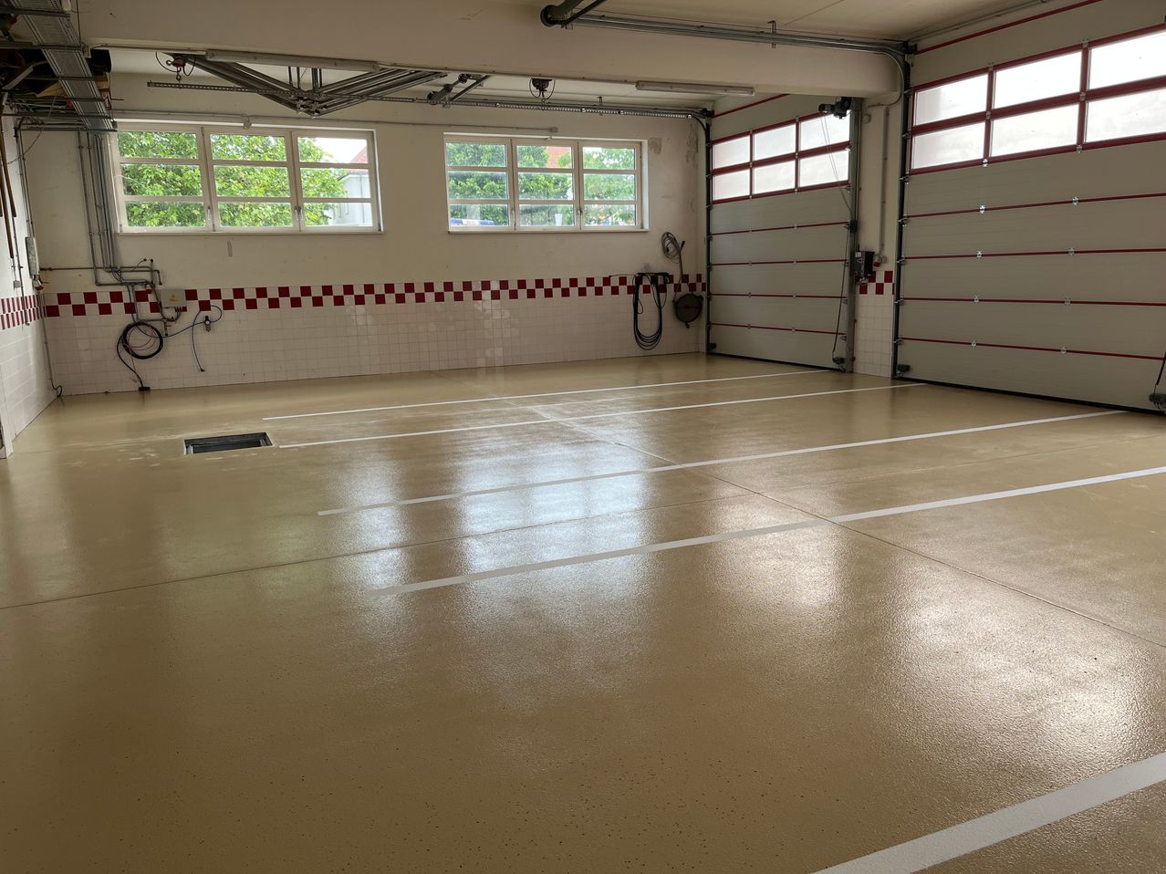 Eine leere Garage mit weiß-rot karierten Wänden, zwei Garagentoren und Fenstern mit Bäumen draußen. Der Boden ist sauber und gepflegt.