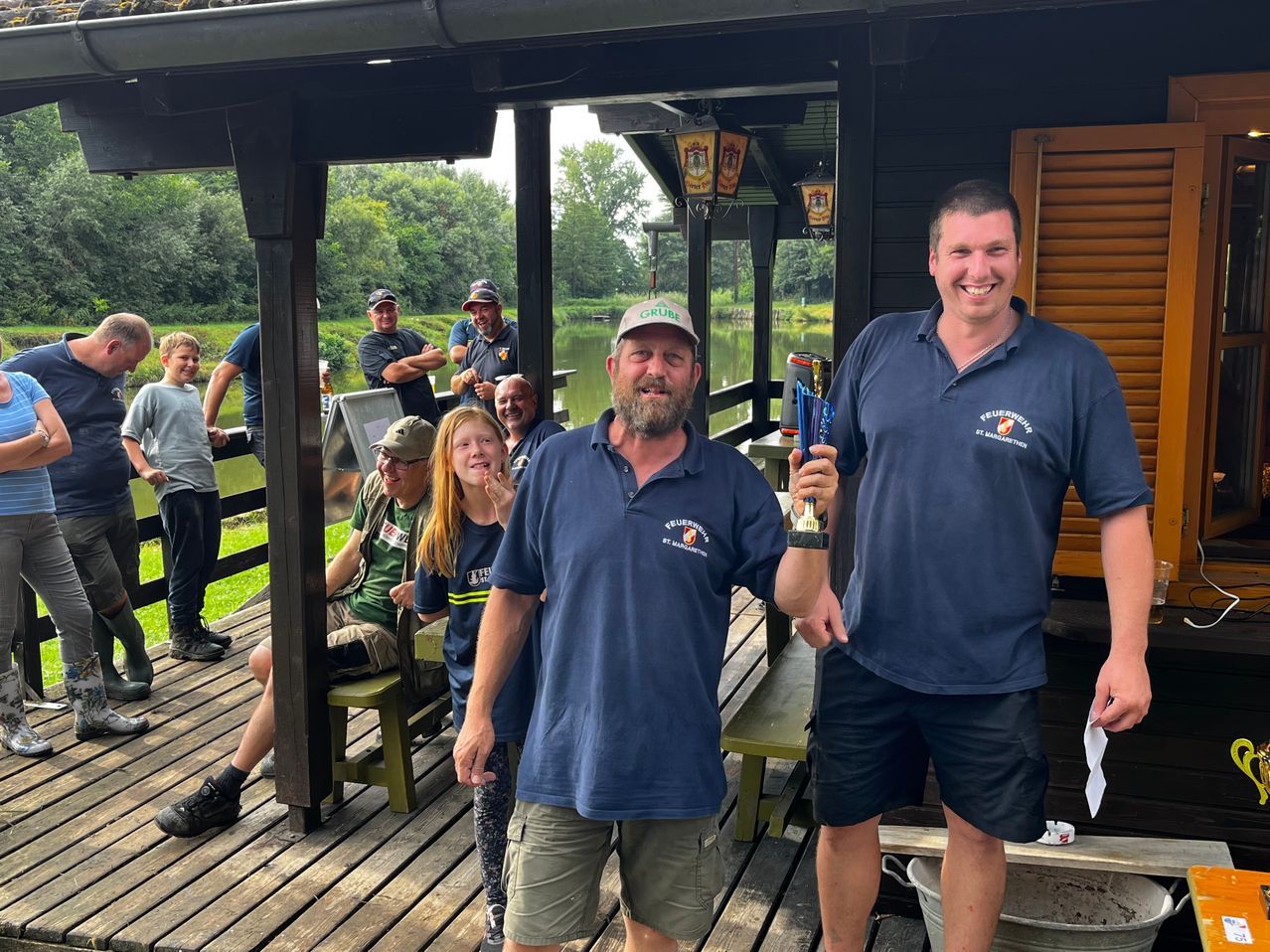 Eine Gruppe von Menschen ist auf einer Veranda versammelt, zwei Männer halten eine Trophäe und lächeln, und andere beobachten sie.