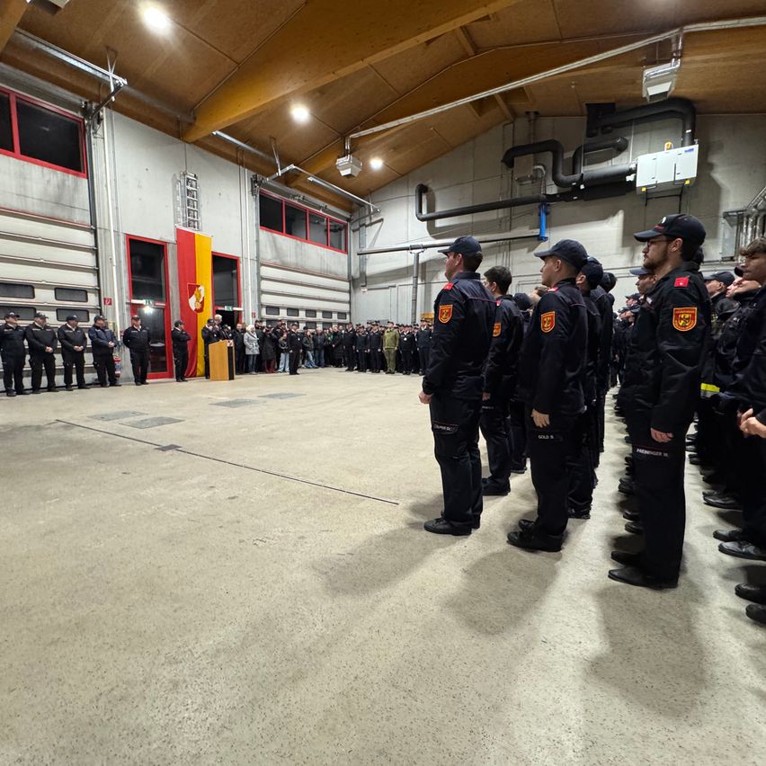 Eine Gruppe von Feuerwehrleuten in Uniform steht in einem Raum mit einer Flagge und einem Podium. Eine Menschenmenge ist hinter ihnen versammelt.