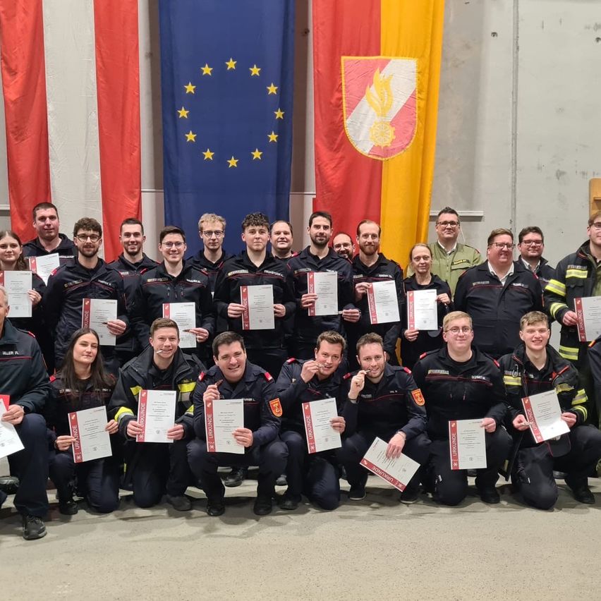 Eine Gruppe von Feuerwehrleuten und Offizieren posiert mit Urkunden vor Fahnen.