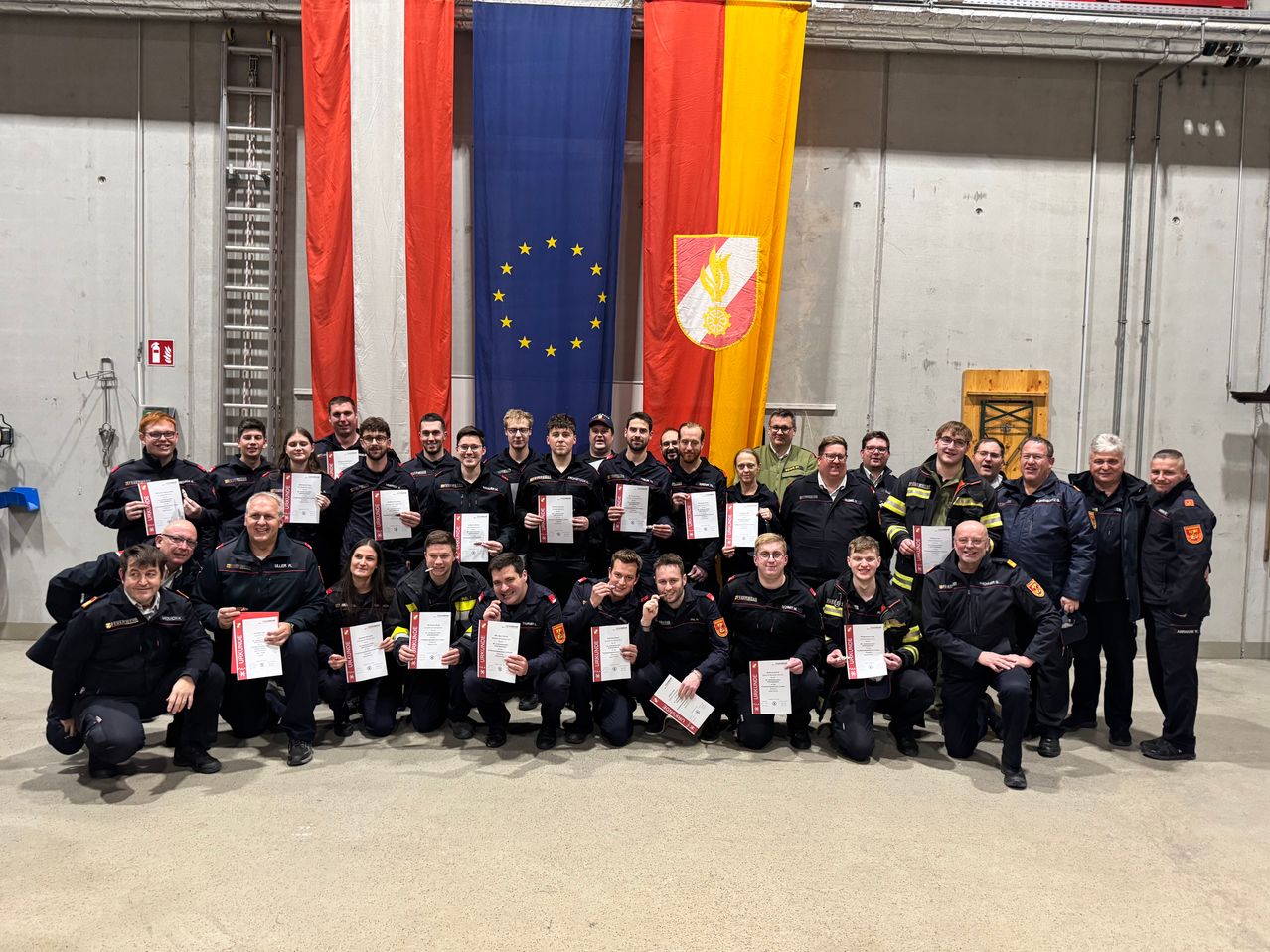 Eine Gruppe Feuerwehrleute in Uniform posiert mit Zertifikaten vor Flaggen der Europäischen Union und einem Wappen.