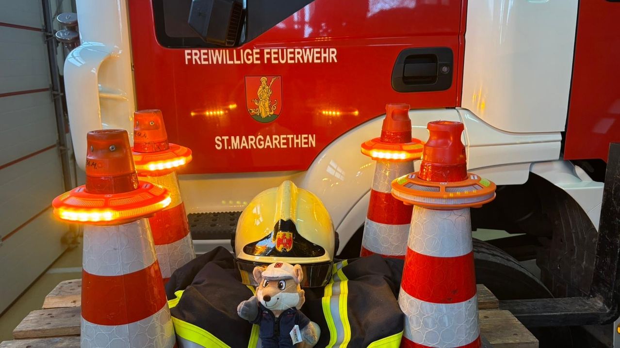 Ein Feuerwehrhelm und eine Feuerwehrjacke liegen auf einem Fahrzeug. Um das Fahrzeug herum befinden sich Kegel und Notfallleuchten. Das Fahrzeug trägt die Aufschrift 'Freiwillige Feuerwehr' und 'St. Margarethen'.