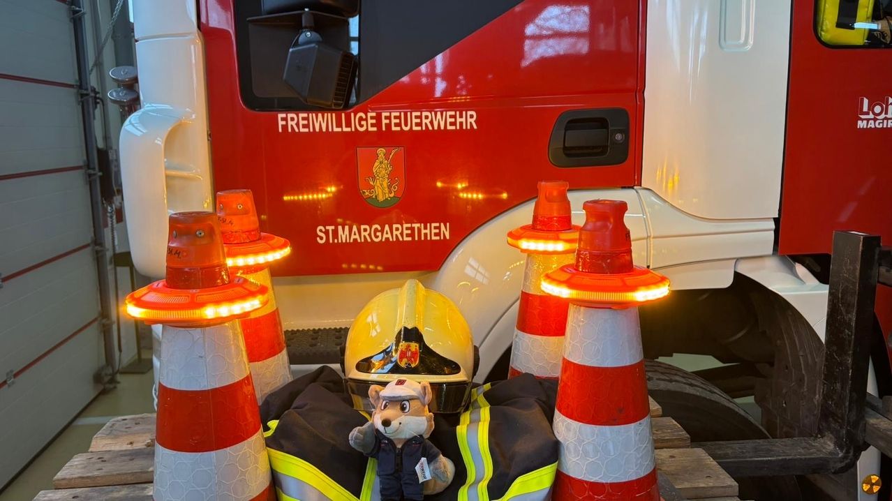 Ein Feuerwehrwagen mit der Aufschrift Freiwillige Feuerwehr St. Margarethen. Davor stehen mehrere orangefarbene Verkehrskegel mit Lichtern. Ein in einem Feuerwehruniform gekleidetes Kuscheltier liegt auf einem Feuerwehrmantel.