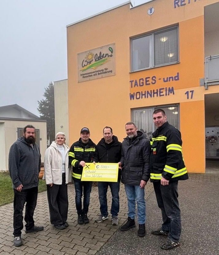 Fünf Personen stehen vor einem gelben Gebäude mit einem Schild, auf dem 'Wir leben' und 'Tages- und wohnheim 17' steht. Ein Mann hält ein gelbes Papier mit dem Text 'C 500-' und einem Kreuzsymbol.