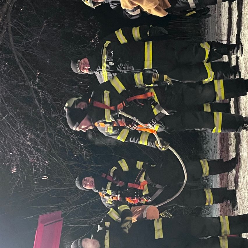 Eine Gruppe von Feuerwehrleuten in voller Ausrüstung, einschließlich Helme und Gurte, versammelt sich nachts im Freien. Einige halten eine aufgerollte Feuerwehrschläuche.