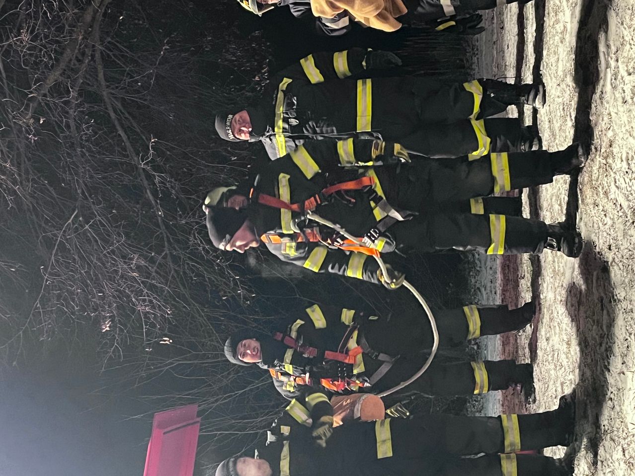 Eine Gruppe von Feuerwehrleuten in voller Ausrüstung, einschließlich Helme und Gurte, versammelt sich nachts im Freien. Einige halten eine aufgerollte Feuerwehrschläuche.