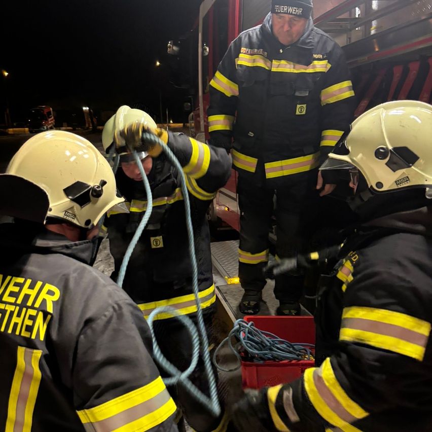 Vier Feuerwehrleute in gelben und schwarzen Uniformen bereiten Ausrüstung vor. Ein Feuerwehrmann hält ein Seil, während andere neben ihm stehen. Eine Aufbewahrungsbox steht auf der Leiter.