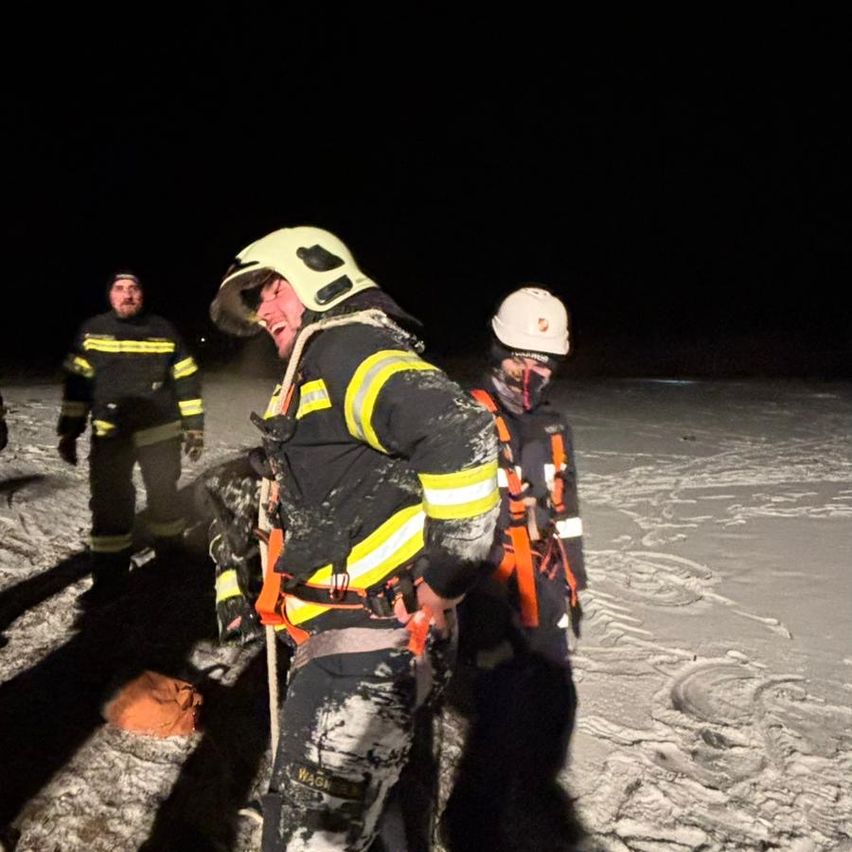 Drei Feuerwehrleute in Schutzkleidung stehen bei Nacht in einem verschneiten Bereich. Einer lacht, ein anderer schaut nach unten und der dritte ist im Hintergrund.