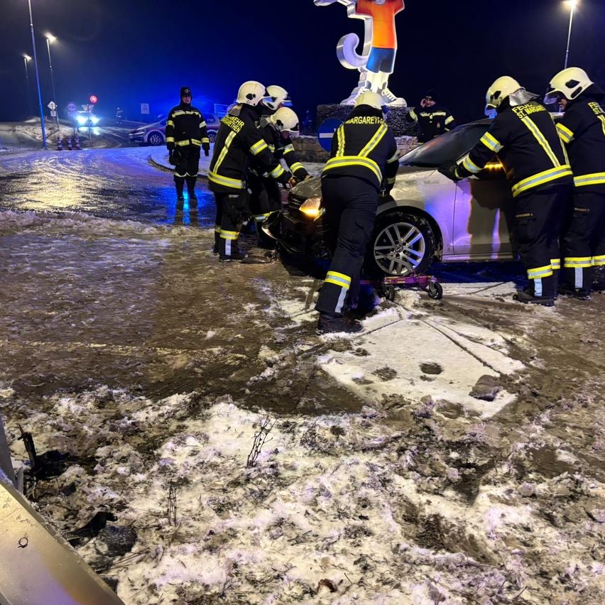 Feuerwehrleute kümmern sich bei Nacht auf einer verschneiten Straße um einen Autounfall, mit einer großen Statue im Hintergrund.