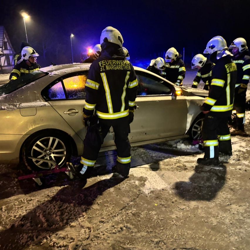 Feuerwehrleute helfen einem festgefahrenen Auto auf einer verschneiten Straße bei Nacht. Sie tragen Helme und Uniformen.