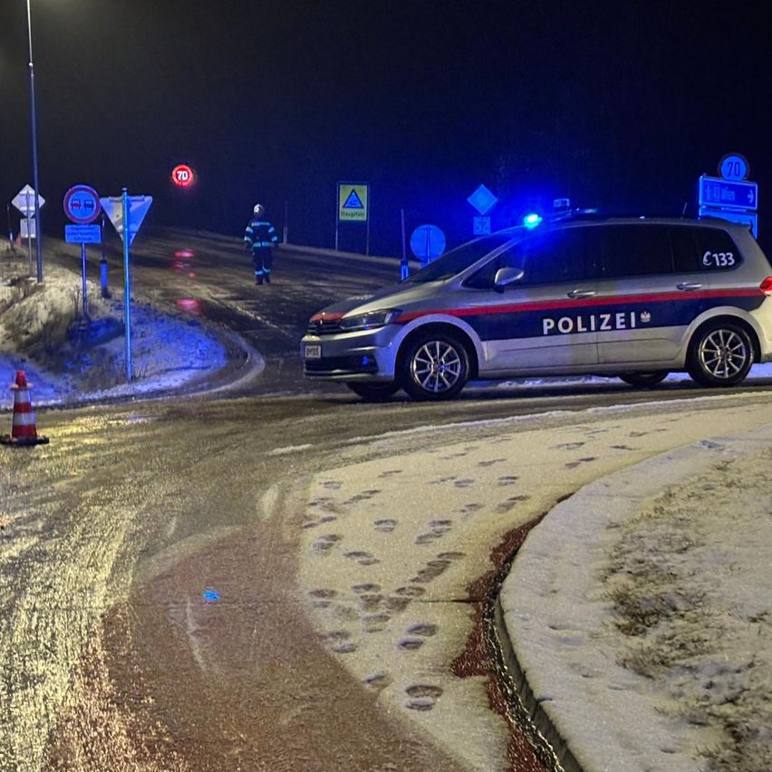 Ein Polizeiauto ist nachts auf einer verschneiten Straße geparkt, mit Fußabdrücken im Schnee. Eine Person in einer Warnweste und Helm geht auf der Straße. Mehrere Verkehrsschilder und ein Verkehrskegel befinden sich am Straßenrand.