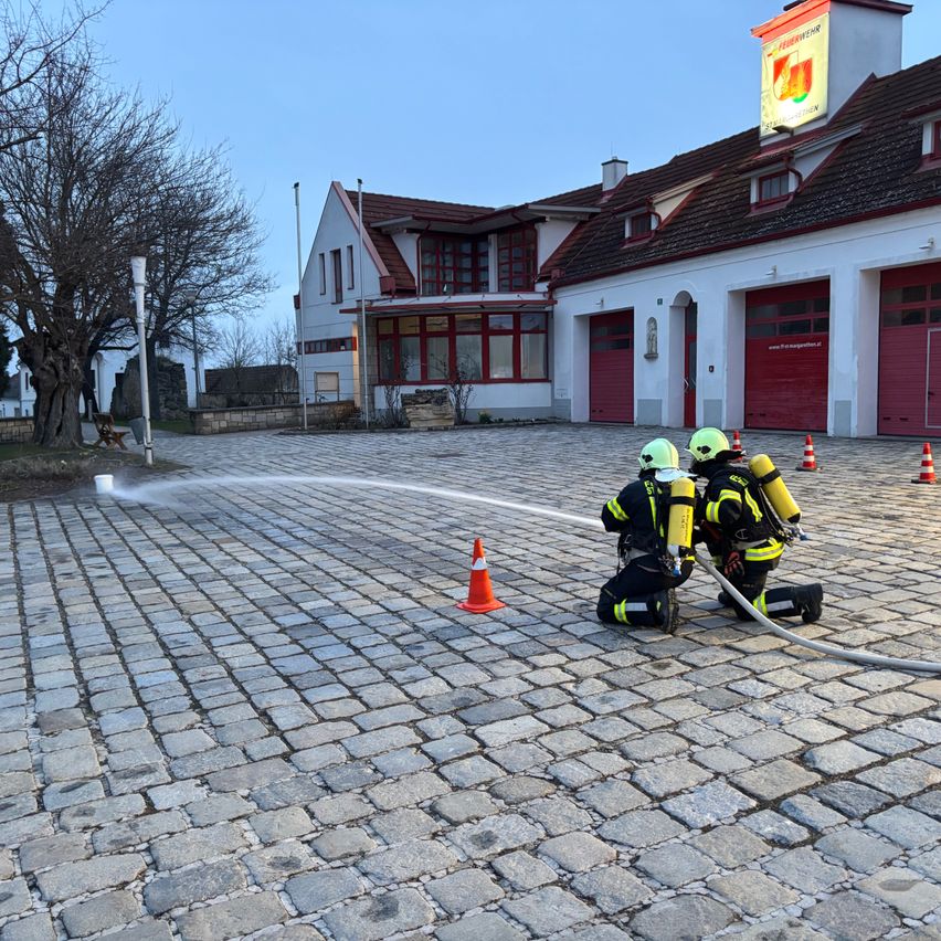 Zwei Feuerwehrleute üben mit einem Feuerwehrschlauch vor einer Feuerwehrstation mit roten Türen.