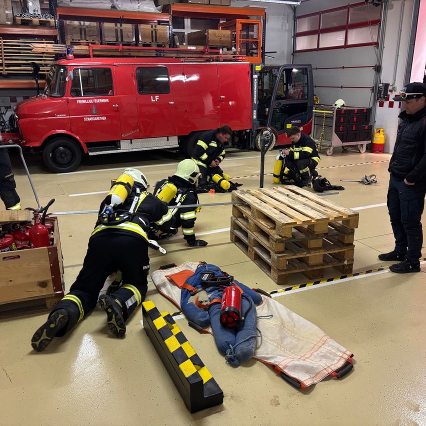 Feuerwehrleute uben mit einer Puppe am Boden. Gerate und Werkzeuge sind in der Nahe, und ein roter Lastwagen ist im Hintergrund.