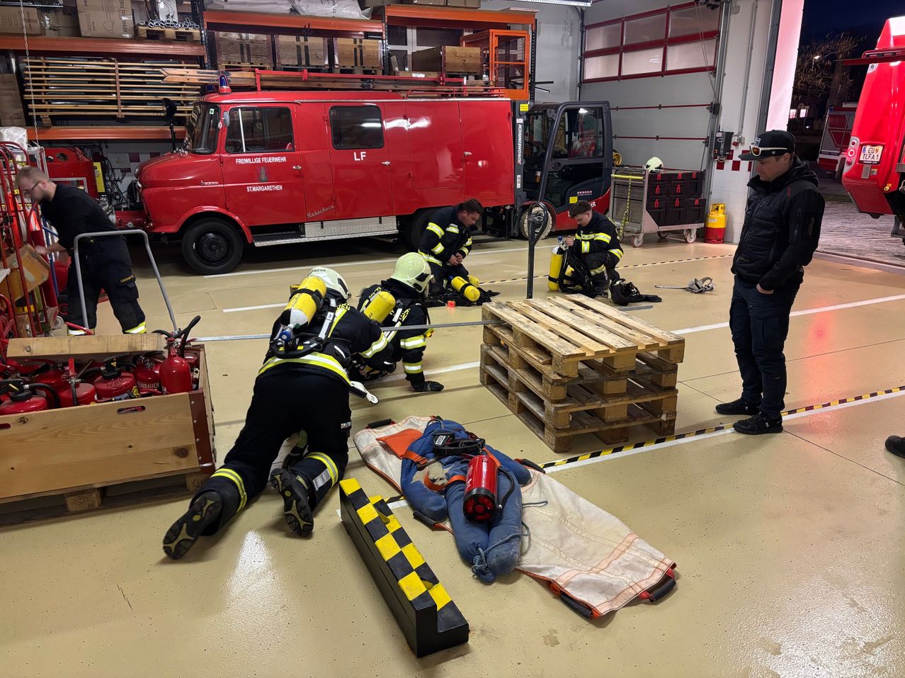 Feuerwehrleute uben mit einer Puppe am Boden. Gerate und Werkzeuge sind in der Nahe, und ein roter Lastwagen ist im Hintergrund.