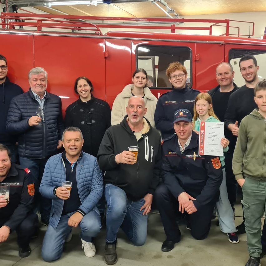 Eine Gruppe von Menschen, darunter Feuerwehrleute, posiert für ein Foto vor einem Feuerwehrwagen und hält Bierbecher in der Hand. Ein Mann hält ein Zertifikat.