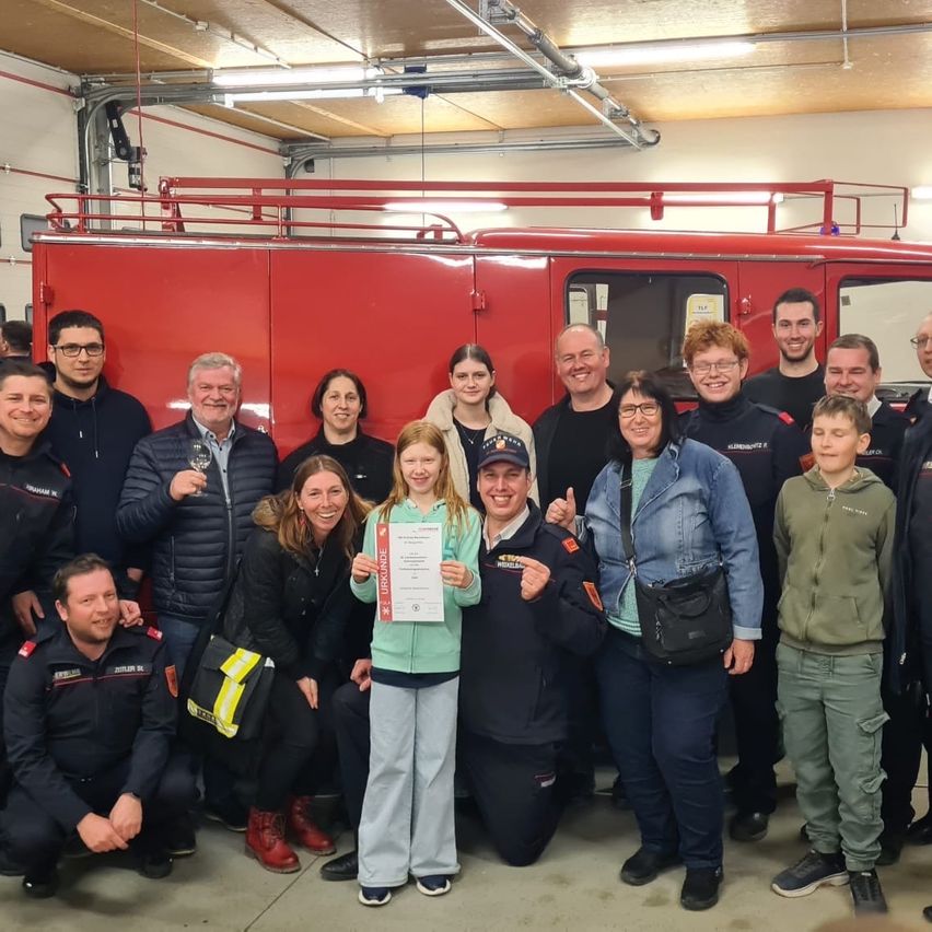 Eine Gruppe von Feuerwehrleuten und Freiwilligen vor einem Feuerwehrauto. Ein junges Mädchen hält ein Zertifikat, während andere lächeln und für ein Foto posieren.