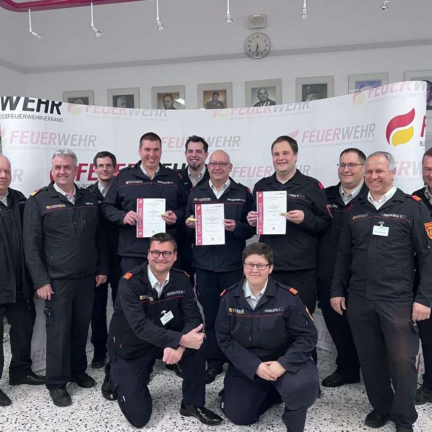 Eine Gruppe von Feuerwehrleuten posiert für ein Foto. Sie halten Urkunden in den Händen und stehen vor einem Banner mit dem Wort Feuerwehr.