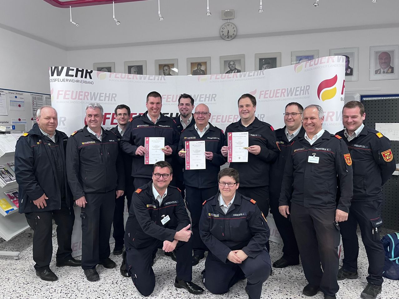 Eine Gruppe von Feuerwehrleuten posiert für ein Foto. Sie halten Urkunden in den Händen und stehen vor einem Banner mit dem Wort Feuerwehr.
