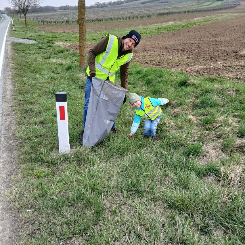 Ein Mann und ein Kind in Warnwesten sammeln neben einer Straße mit einem Pfahl und einem Baum Müll auf.