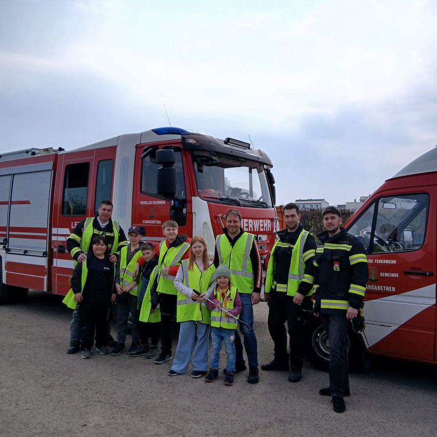 Eine Gruppe von Kindern und Erwachsenen in Warnwesten posiert vor zwei Feuerwehrwagen an einem bewölkten Tag.