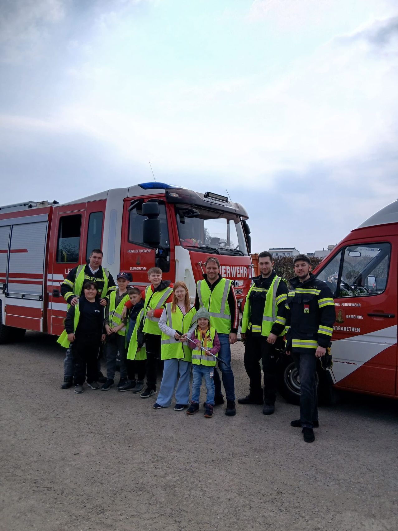 Eine Gruppe von Kindern und Erwachsenen in Warnwesten posiert vor zwei Feuerwehrwagen an einem bewölkten Tag.