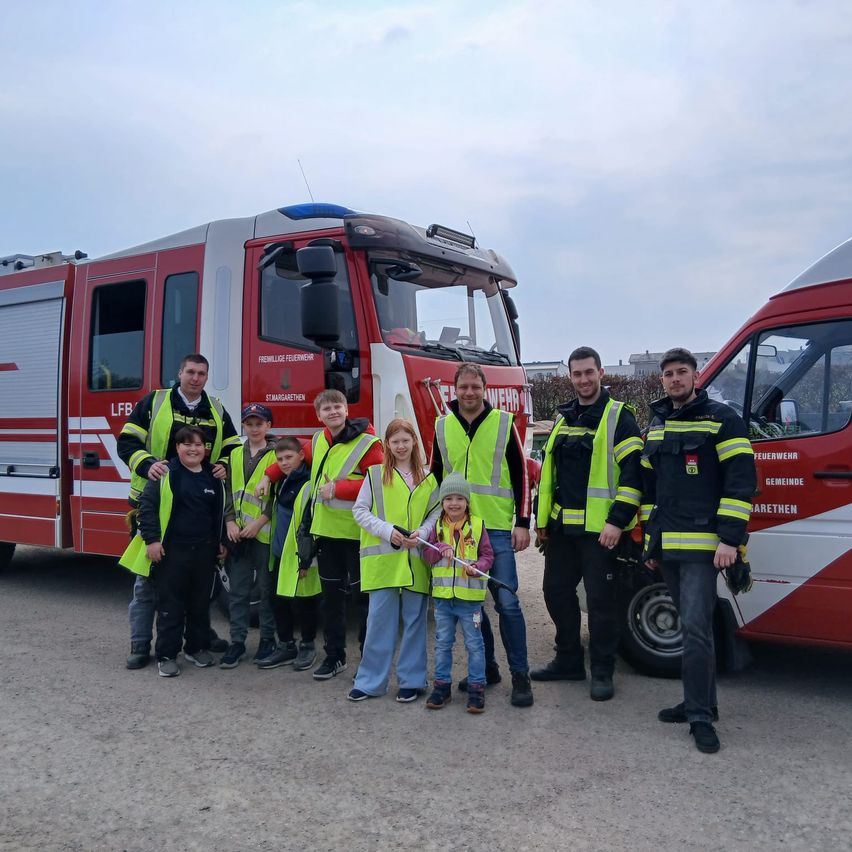 Eine Gruppe von Erwachsenen und Kindern in gelben Warnwesten posiert für ein Foto vor zwei Feuerwehrfahrzeugen.