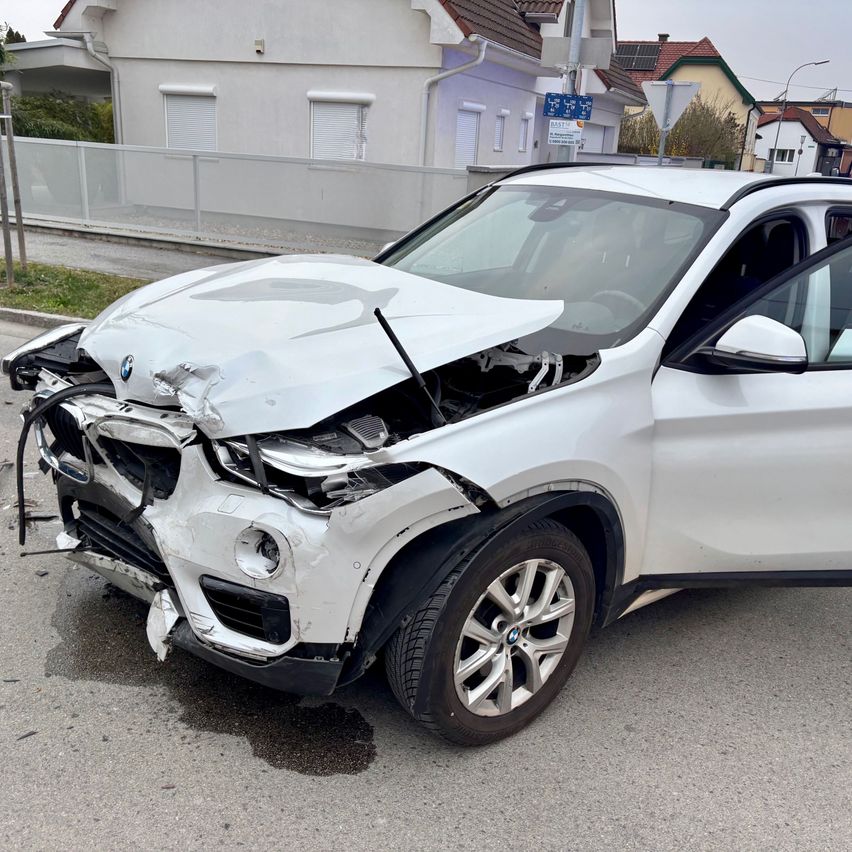 Ein weißer BMW X3 ist mit stark beschädigter Front auf der Straße geparkt. Der Stoßfänger ist verbogen und die Scheinwerfer sind zertrümmert. Die Motorhaube des Autos ist geöffnet.
