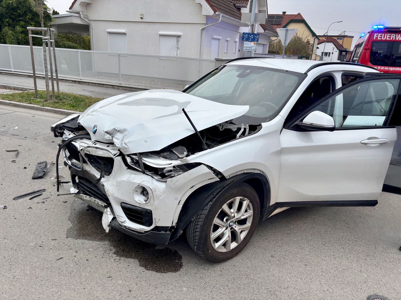 Ein weißer BMW X3 ist mit stark beschädigter Front auf der Straße geparkt. Der Stoßfänger ist verbogen und die Scheinwerfer sind zertrümmert. Die Motorhaube des Autos ist geöffnet.