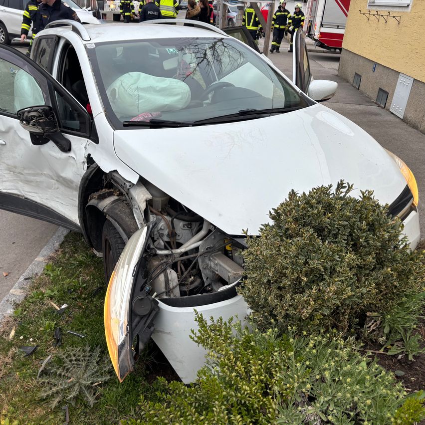 Ein weißer Wagen ist in einen Busch auf der Straßenseite gecrasht. Das Auto hat schwere Schäden am vorderen Stoßfänger und der Motorhaube. Rettungsdienste sind anwesend.