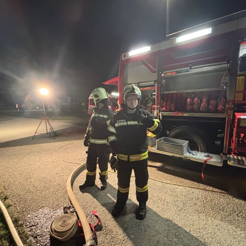 Zwei Feuerwehrleute in voller Ausrüstung stehen neben einem Feuerwehrauto in der Nacht, mit Ausrüstung und einem Schlauch in der Nähe.