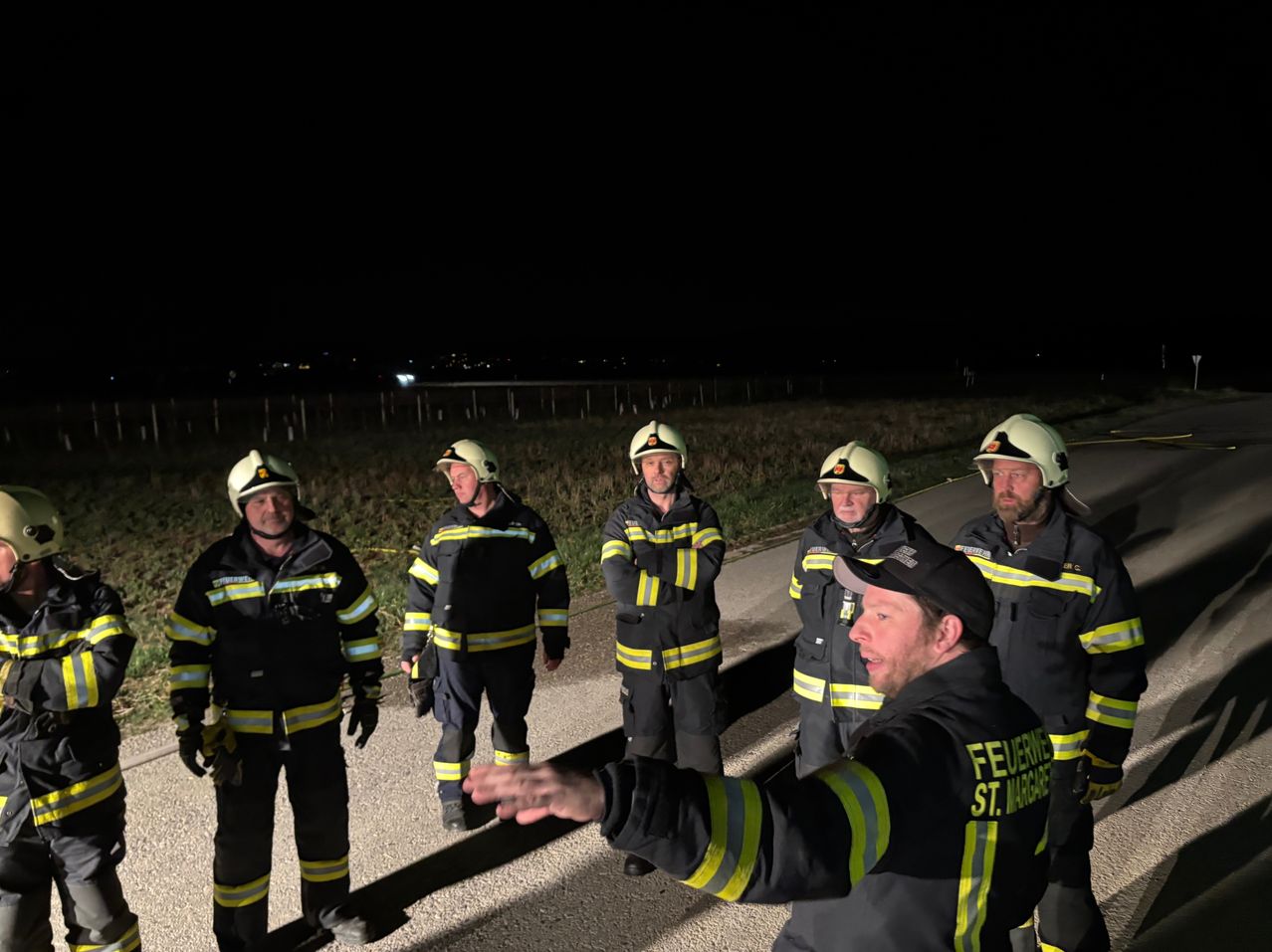 Fünf Feuerwehrleute stehen nachts auf einer Straße; einer zeigt auf etwas, und die anderen hören zu. Der Hintergrund zeigt ein Grasfeld und eine Stadtsilhouette.