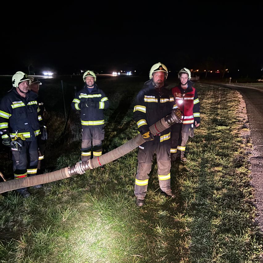 Fünf Feuerwehrleute stehen nachts draußen, einer hält eine lange Schlauch und eine Taschenlampe. Sie tragen gelbe und schwarze Uniformen mit Helmen und Handschuhen.