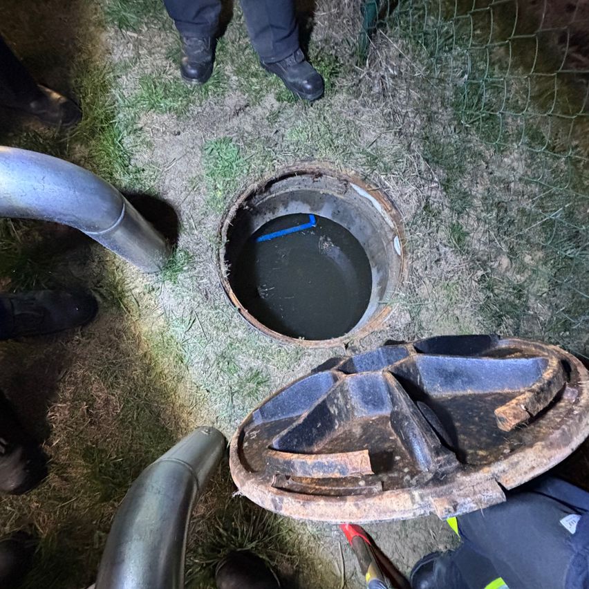 Offener Schacht auf Gras mit einer blauen Linie im Inneren, umgeben von Menschen in schwarzen Schuhen. Ein rostiges Metalldeck liegt in der Nähe.