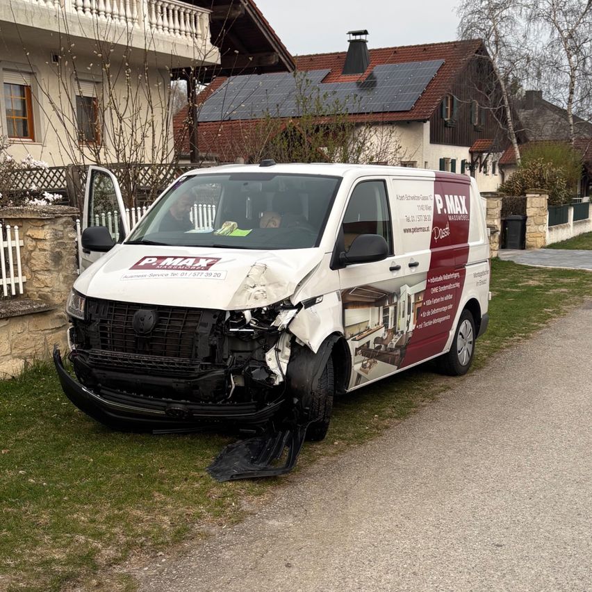 Ein beschädigter weißer Van mit rotem und weißem Design steht mit beschädigtem vorderen Stoßfänger auf einer Einfahrt. Hinter dem Van steht ein Haus mit Solarmodulen.
